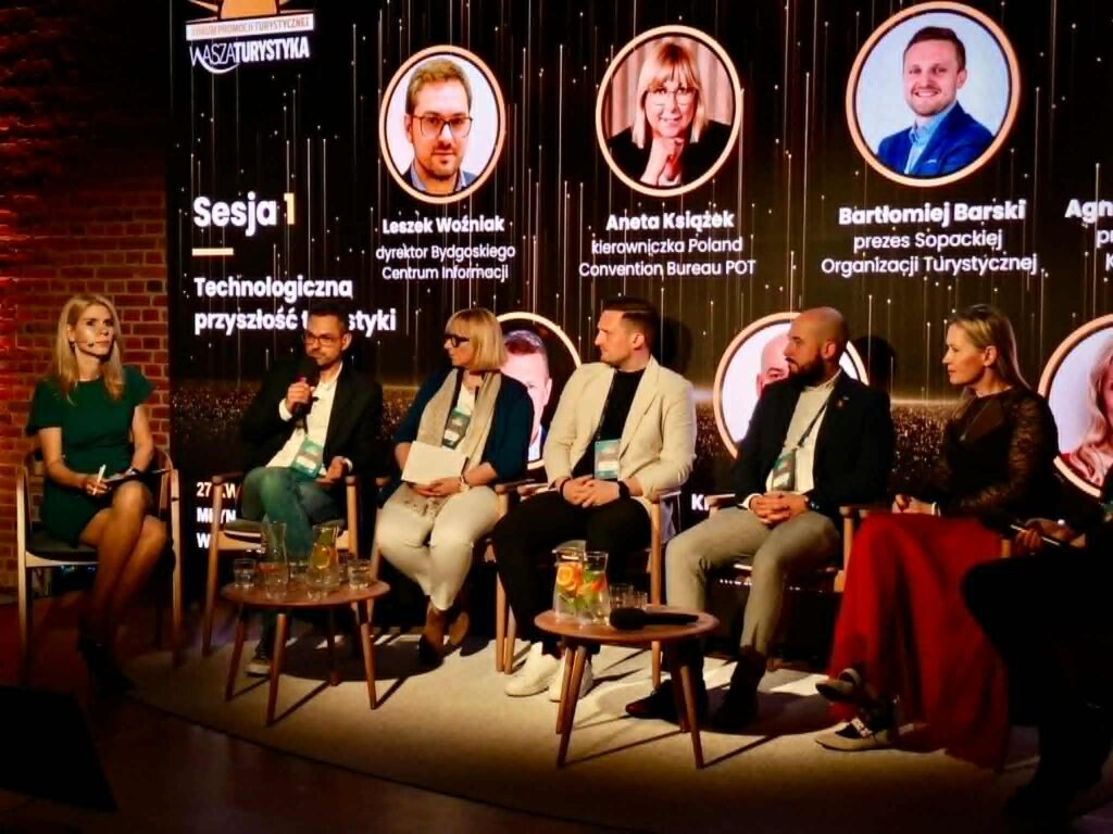 Sesja 1, na scenie siedzi 7 osób i prowadzą dyskusję na temat technologicznej przyszłości turystyki